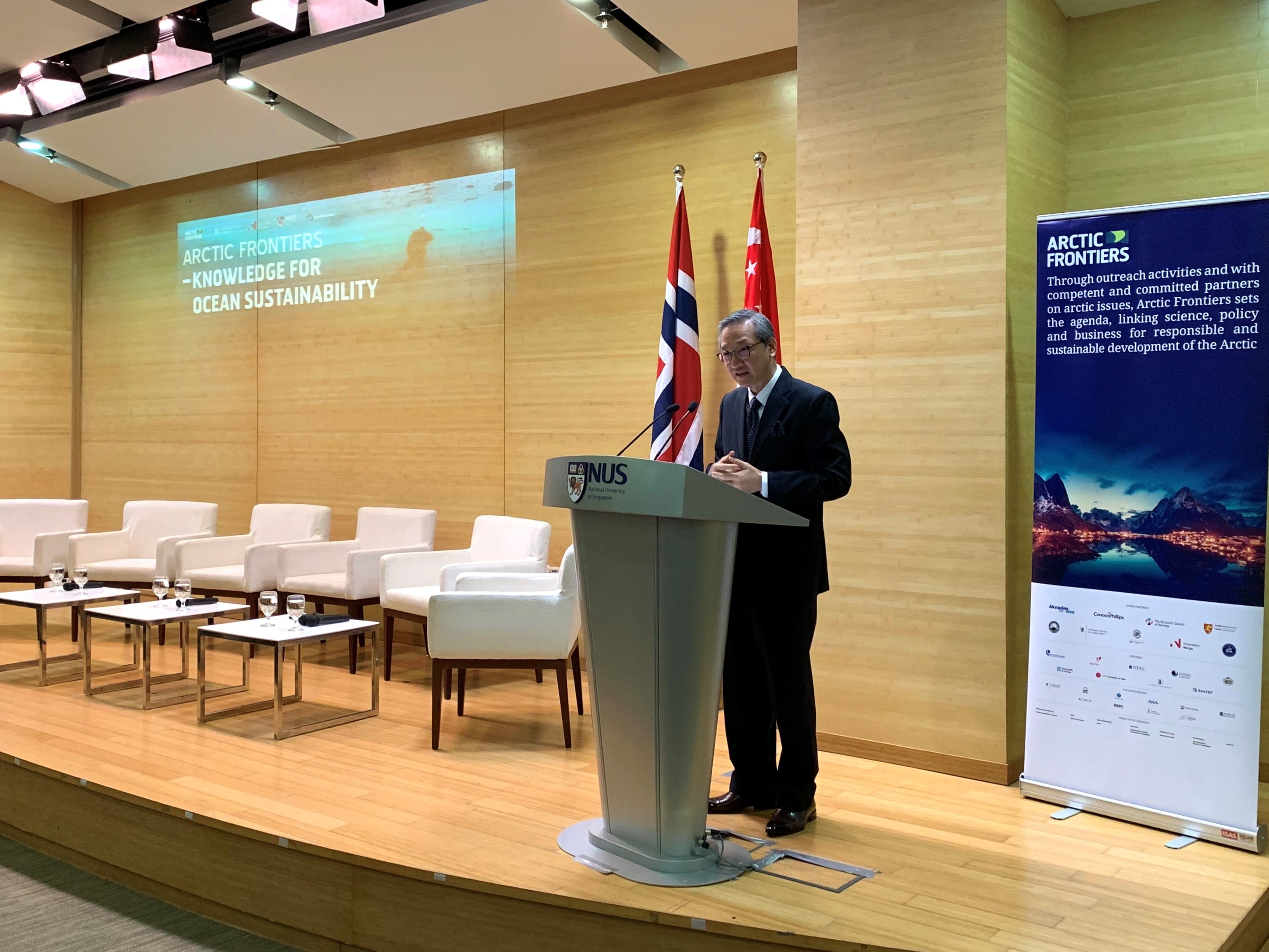 Speaker at NUS lectern by Norway & Singapore flags; "Arctic Frontiers" banner & screen visible.